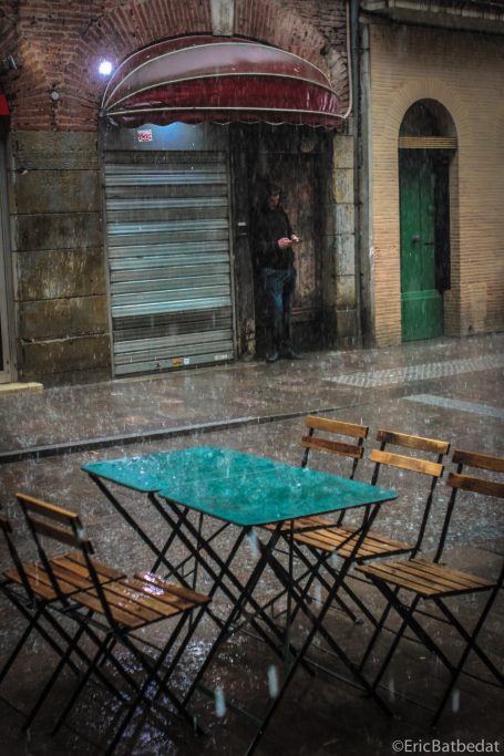 Table et chaises mouillées sous la pluie, devant une entrée de café fermée.