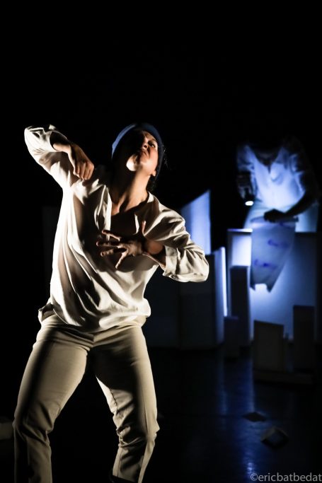 Danseur en mouvement sur scène, éclairage sombre avec des décors abstraits en arrière-plan.