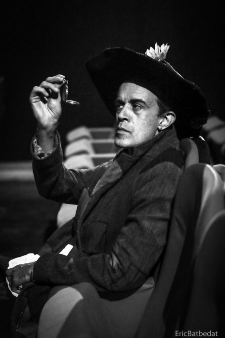 Homme en costume sombre et chapeau, tenant une cigarette, assis dans un théâtre.