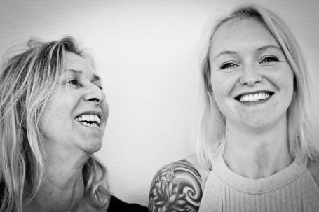 Deux femmes souriantes, l'une avec des cheveux blonds et l'autre avec un tatouage à l'épaule.