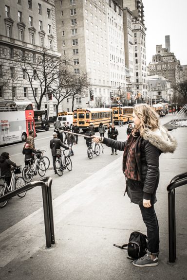 Femme en manteau tenant un smartphone, avec des cyclistes et des bus en arrière-plan.