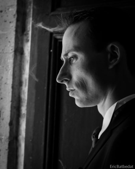 Homme en costume noir regardant à travers une fenêtre, expression pensive.