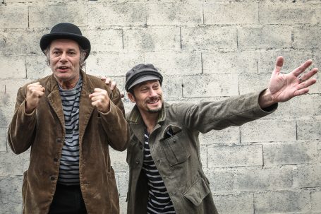Deux hommes en costume rayé, posant joyeusement devant un mur gris.