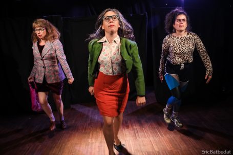 Trois femmes en tenues variées marchent avec assurance sur scène, éclairées par des projecteurs.