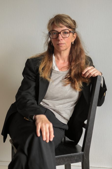 Femme assise sur une chaise, portant des lunettes et un costume noir.