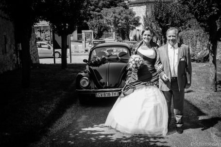 Femme en robe de mariée avec un homme, devant une voiture rétro sur une allée.