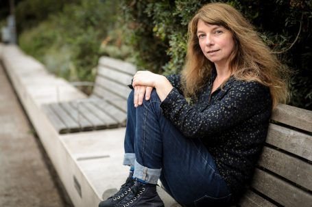 Femme assise sur un banc, regard pensif, cheveux longs et ondulés, vêtements décontractés.