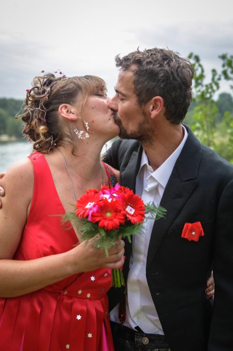 Un couple romantique échange un baiser, la femme portant une robe rouge et un bouquet de fleurs.