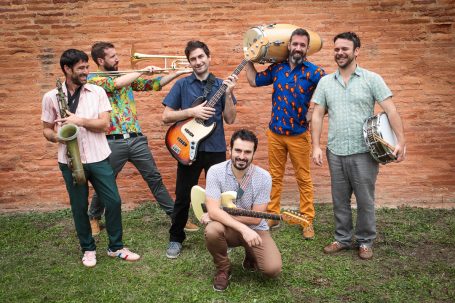 Groupe de musique composé de six membres, souriants, sous un mur de briques.