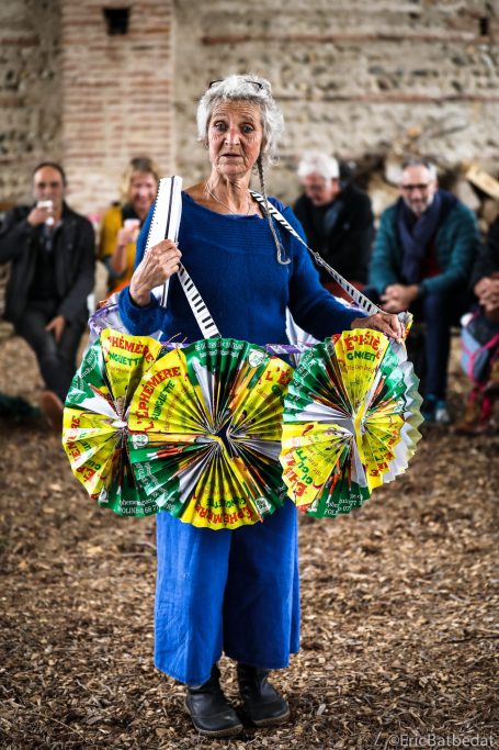 Femme souriante en tenue bleue portant des paniers colorés, entourée de personnes.