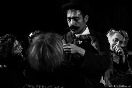 Un homme en costume réalise une performance théâtrale en noir et blanc devant un public attentif.