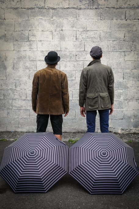 Deux hommes de dos, portant des chapeaux, devant un mur, avec deux parapluies rayés à leurs côtés.