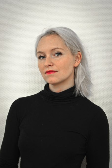 Femme aux cheveux blonds platine, portant un haut noir, avec un rouge à lèvres rouge.