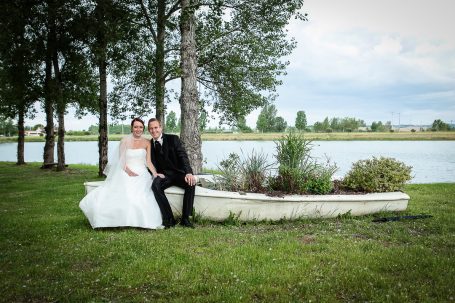 Un couple de mariés assis sur un banc près d'un plan d'eau, entouré d'arbres.